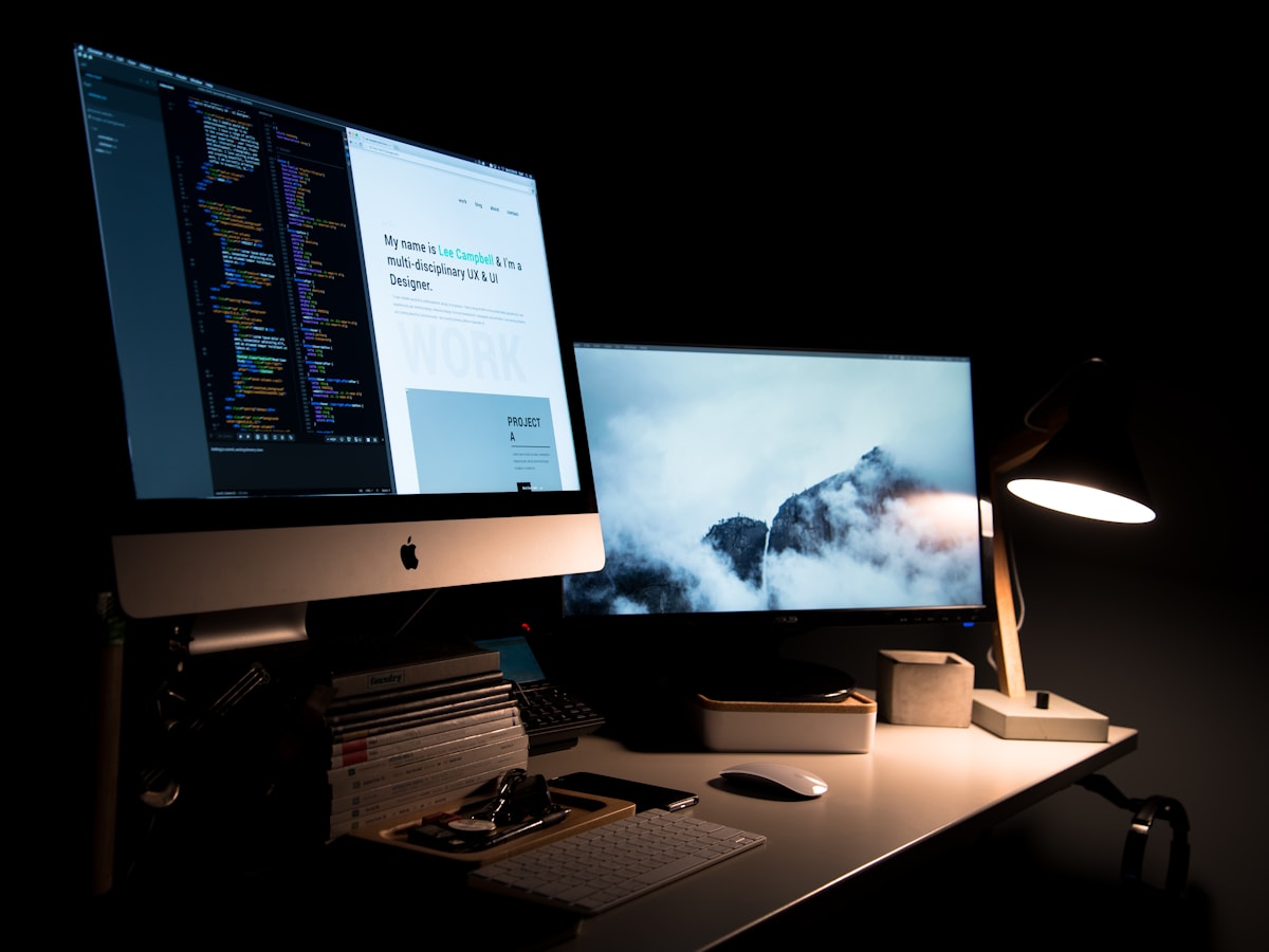 A designer's dark desk setup — code and a portfolio site on an iMac beside a second monitor showing a website